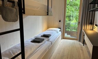 A modern bedroom with a bunk bed and a large window that overlooks a green balcony view. The walls are made of light wood, and the room is simply and invitingly furnished.