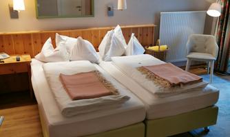 A cozy hotel room with two beds, bright bed covers, and a modern armchair. The walls are painted in neutral colors, and there is a small table.