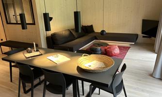 A modern dining room with a black table and chairs. In the background, there is a cozy seating area and large windows provide plenty of light.