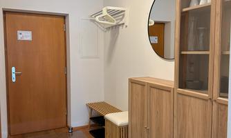 A bright entrance area with a wooden table and a coat rack. A round mirror hangs on the wall and a door leads outside.
