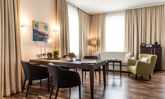 A modern living space with a large table and several chairs. The room has light curtains and a cozy corner with a green armchair.
