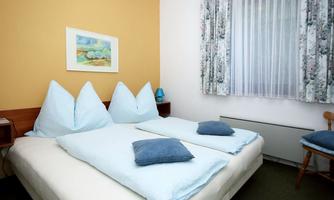 A cozy bedroom with two beds and blue pillows. The walls are in soft colors and there is a lovely curtain.