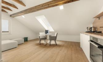 A modern attic apartment with a wooden ceiling and bright walls. The space is minimally furnished with a dining table and a minimalist kitchen.