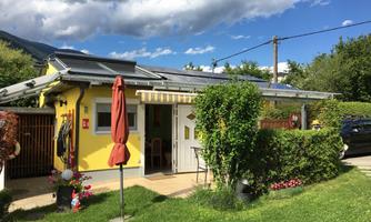 A small, yellow house with a green garden and sun umbrellas. The facade is friendly and inviting, surrounded by plants and trees.