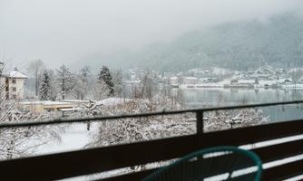 A wintry view of a snowy landscape with a lake and snow-covered trees. In the background, mountains and small houses can be seen.