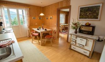 A cozy kitchen with wooden walls and a table with chairs. In the background, there are windows with a view of the outdoors and a vintage sideboard.