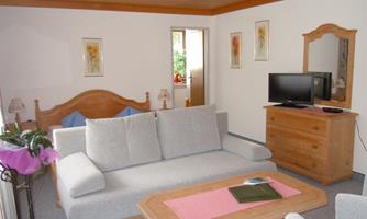 A cozy living room with a gray sofa, a wooden table, and a television. The walls are decorated with floral prints and there is a connection to a bedroom.