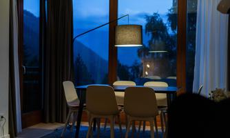 A cozy dining area with a table and white chairs. The large windows provide a view of a gentle evening twilight and the surrounding nature.
