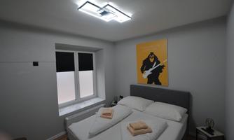 A modern bedroom with a double bed and white bedding. A colorful piece of artwork hangs on the wall, and there is a window with blinds.