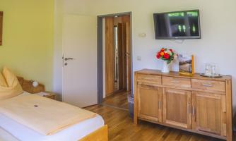 A cozy room with a double bed, wooden floor, and soft colors. A television hangs on the wall, and a dresser with flowers stands beside it.
