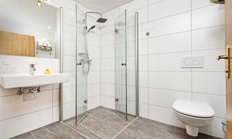 A modern bathroom with a glass shower and a simple sink. The walls are tiled in light colors and the floor has a gray tone.