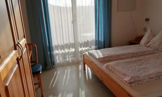 A simple room with a double bed, a chair, and a wardrobe. The light shines through the curtains, creating a bright atmosphere.