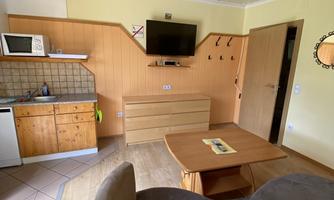 A cozy living space with a small kitchen, a table, and a sofa. On the wall hangs a TV and some hooks.