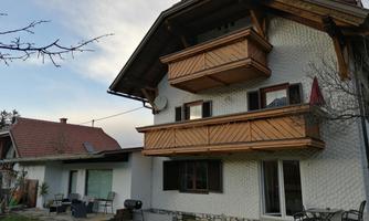 A lovely two-story house with a wooden balcony. In the foreground, there is a garden with seating.