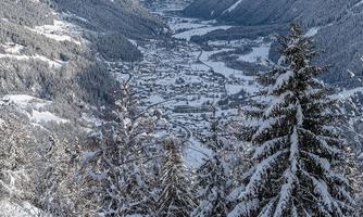A snowy mountain landscape with tall, frost-covered trees. In the valley lies a small village, surrounded by majestic mountains.