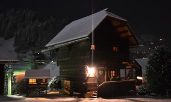 A charming wooden house at night, surrounded by snow. The windows radiate warm light, creating a cozy atmosphere.