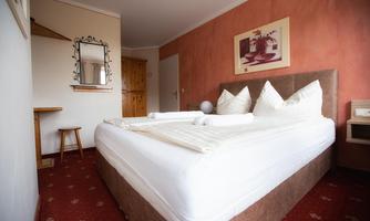 A comfortable bedroom with a large bed and fresh bed linens. The walls are in soft colors, and there is a wooden chair as well as a shelf.
