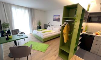 A modern room with a large bed and green accents. There is a small dining area and a kitchenette.