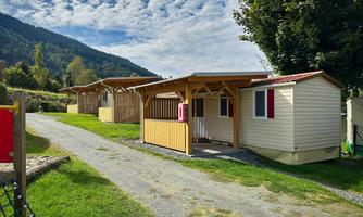 Huts in a campsite, surrounded by a green meadow and mountains in the background. The sky is clear with a few clouds.