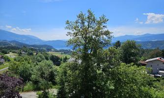 A sunny view of green hills and trees with a clear blue sky. In the background, mountains and a lake can be seen.