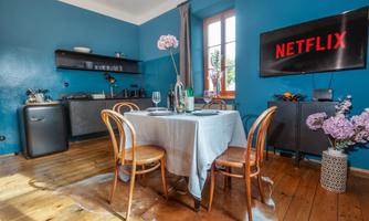 A cozy dining area with a table surrounded by chairs. A television with the Netflix logo hangs on the wall.