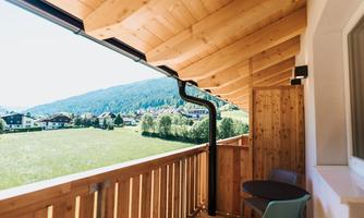 A cozy balcony with wooden cladding and a view of a green meadow and mountains. Two chairs and a table are ready.
