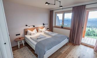 A modern bedroom with a double bed and wooden floor. Large windows provide a view of the landscape.