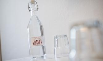 A glass bottle with the label "Krämer" is on a table. Next to it is a glass and another inverted cup.