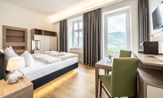 A modern hotel room with a double bed, a desk, and a window that offers a view of the landscape. The decor is bright and cheerful, with wood and green tones.