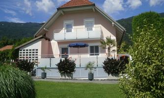 A beautiful pink house with a balcony and garden. In the background, there are green mountains and a blue sky.