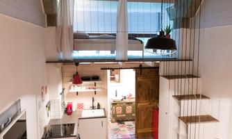 A modern, open living kitchen with wooden shelves and a staircase leading to an upstairs bedroom. The decor is stylish and well-organized, with a cozy atmosphere.