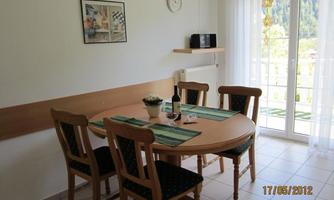 A bright dining room with a round wooden table and four chairs. On the table, there is a plant and a bottle of wine.