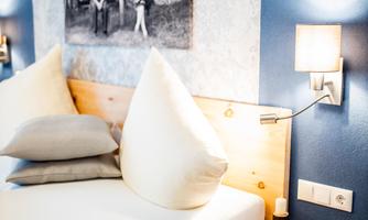 A modern bedroom with a cozy bed, stacked pillows, and a wooden nightstand. On the wall hangs a black and white picture depicting rustic motifs.