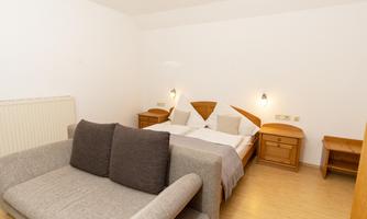 A cozy bedroom with a double bed and a gray couch. The walls are white and there are light wooden furniture.