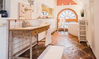 A modern bathroom with a wooden vanity and a large round window niche. The floor consists of rustic tiles and there are simple, bright decorative elements.