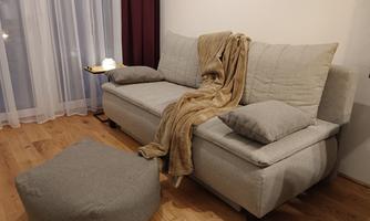 A cozy living room with a grey sofa, an ottoman, and a blanket. The room has large windows with curtains and a small table.