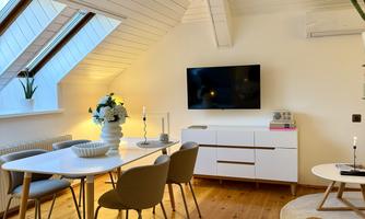 A modern living room with a dining table and chairs. A television is located on the wall, and large windows provide plenty of daylight.
