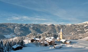 A snowy landscape with traditional cottages and a church. In the background, the snow-covered mountains rise beneath a clear blue sky.
