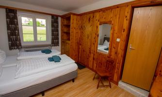 A cozy bedroom with two single beds and wooden furniture. The room has a window with a view of greenery and a small dresser.