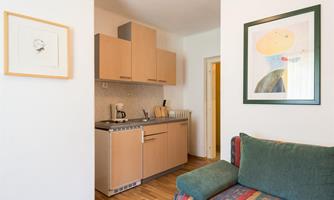 A small, modern kitchen with wooden cabinets and appliances. Next to the kitchen is a cozy sofa with colorful cushions.