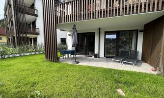 A modern terrace with loungers and a sun umbrella. The garden is well-kept and surrounded by green plants.