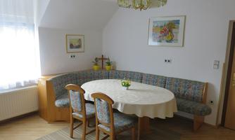 A cozy dining area with a round tablecloth and a large, cushioned seating area. Framed by windows and pictures, the room exudes a welcoming atmosphere.