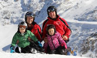 A family sits in the snow on a ski slope. They are wearing ski gear and smiling at the camera, surrounded by snow-covered mountains.