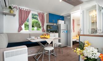 A cozy living area with a table, chairs, and modern kitchen equipment. Large windows provide plenty of daylight and offer a view of the greenery.
