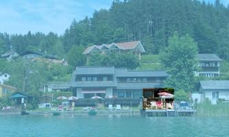 A quiet lakeshore with several houses and trees in the background. On the terrace, sun umbrellas and loungers can be seen.