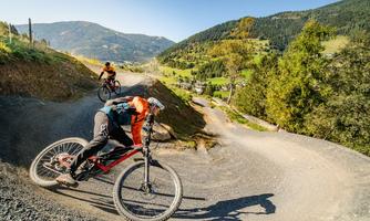 A mountain biker rides quickly down a winding path. In the background, there are green hills and a clear blue sky.