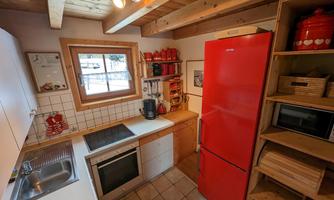 A modern kitchen with red accents and wooden details. The refrigerator is bright red and the windows offer a view of the surroundings.