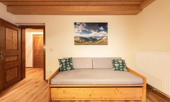 A cozy living room with a wooden sofa and two cushions. A landscape painting hangs on the wall, and the floor is made of wood.