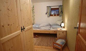 A cozy bedroom with a wooden bed and blue bedding. Next to the bed is a nightstand with a lamp and a small chair.