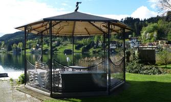 A pavilion by the shore of a lake, surrounded by green grass and trees. In the background, there are pretty houses and a picturesque landscape.